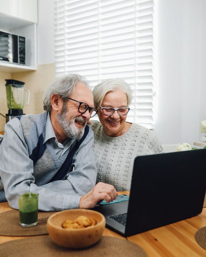 Casal de idosos rindo juntos enquanto usam um notebook na cozinha de casa