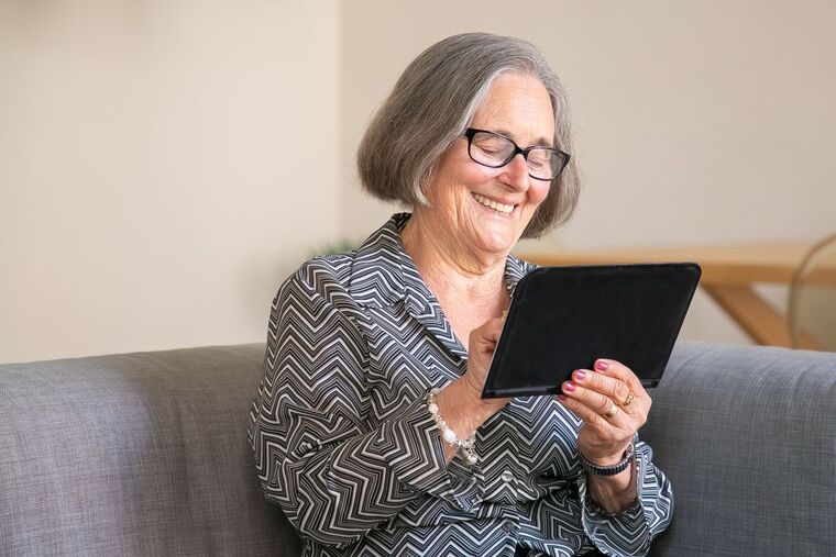 Mulher de cabelos grisalhos sorrindo enquanto usa um tablet no sofá de casa